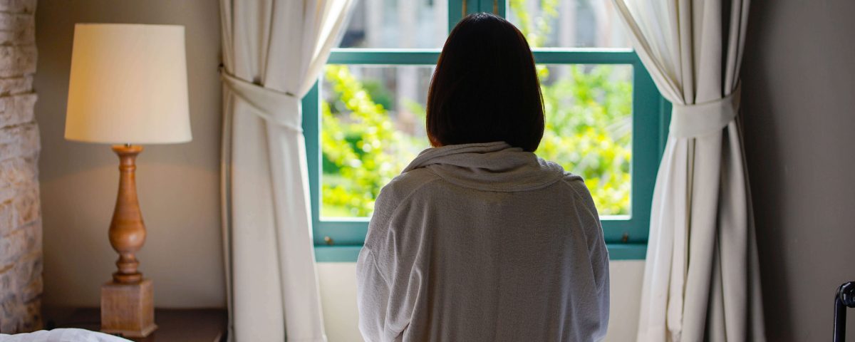Woman with short dark hair sitting on a bed looking out a window
